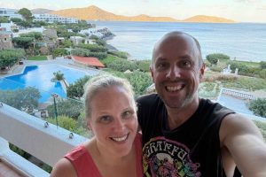 rachel and sasha standing on a hotel room balcony at Elounda Mare hotel in Elounda, Greece with a swimming pool, the ocean, and mountains behind them